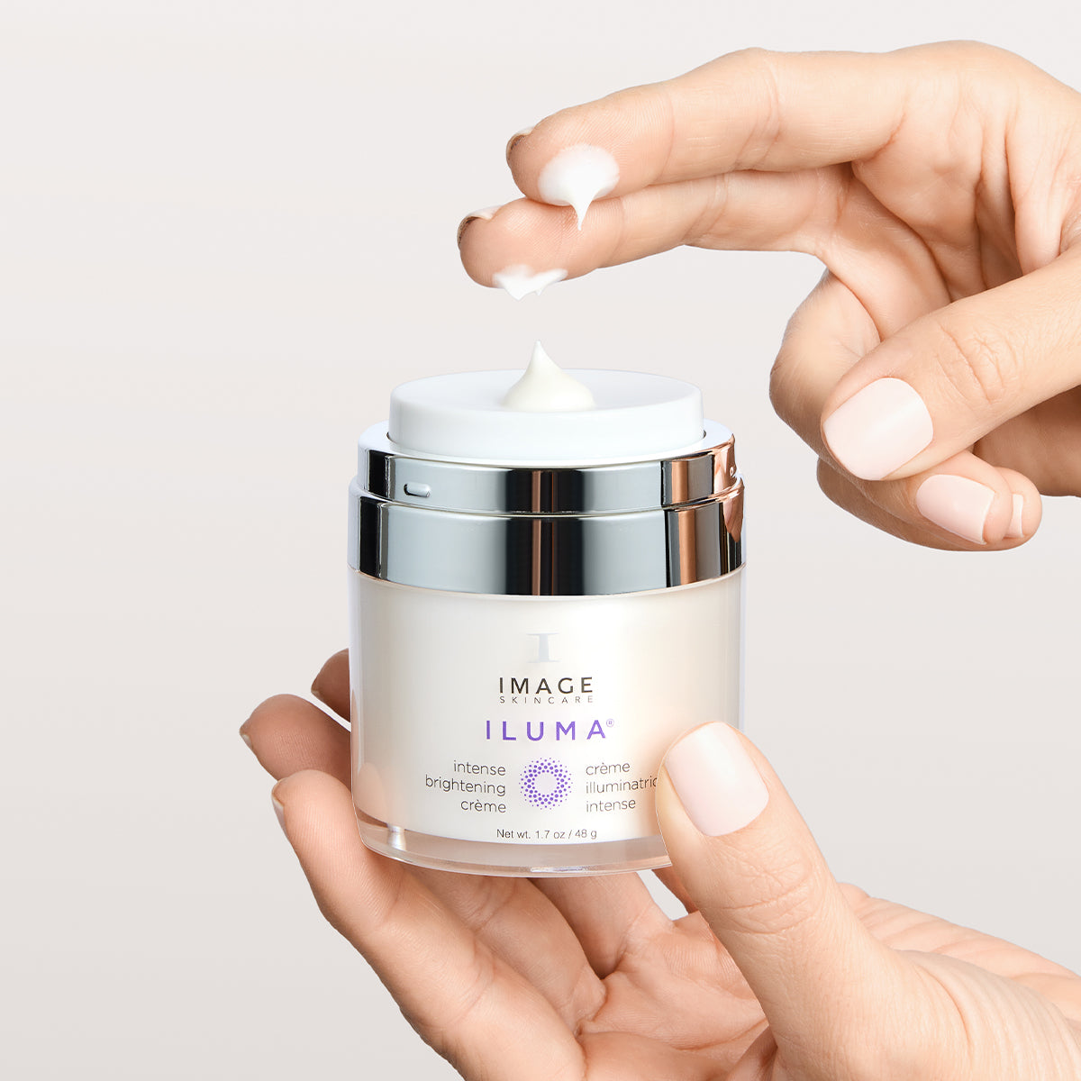 model hands holding iluma brightening creme container with fingers dipped in product